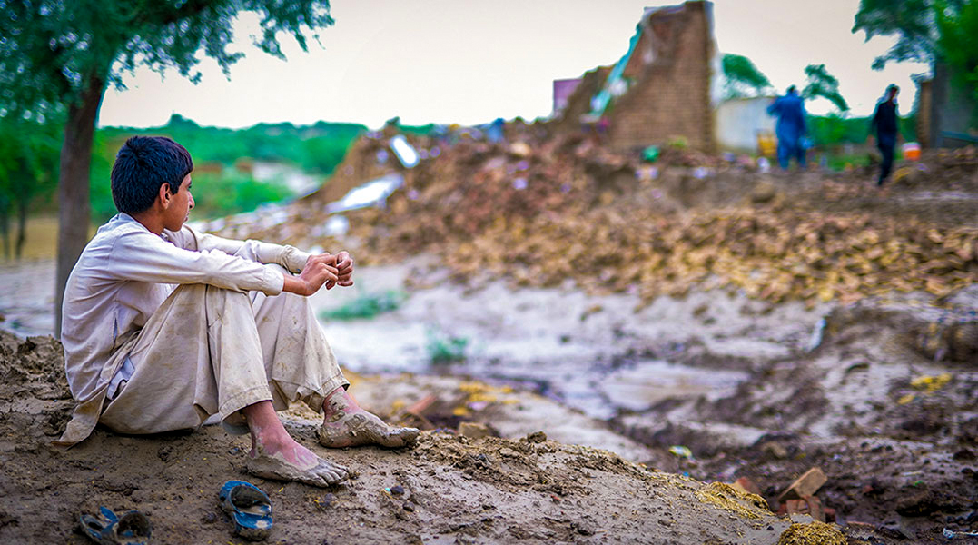 A displaced young man sits alone beside a mud-covered path in front of a collapsed brick house after floodwater has receded
